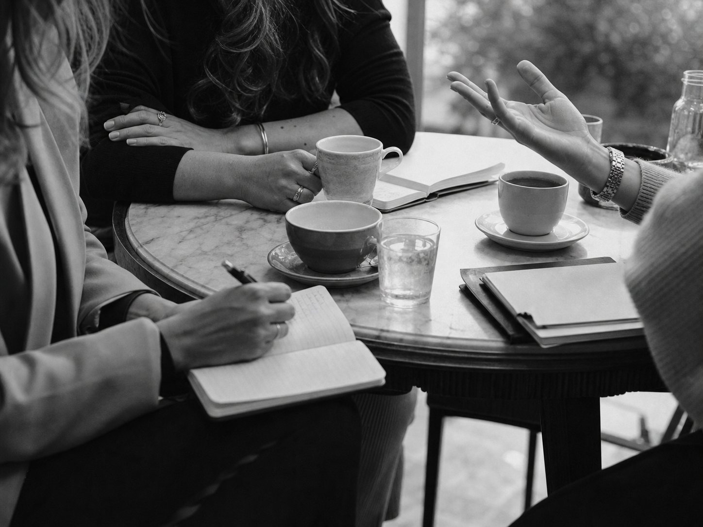 Mulheres em conversa à volta de uma mesa com cadernos e café, sem rostos visíveis